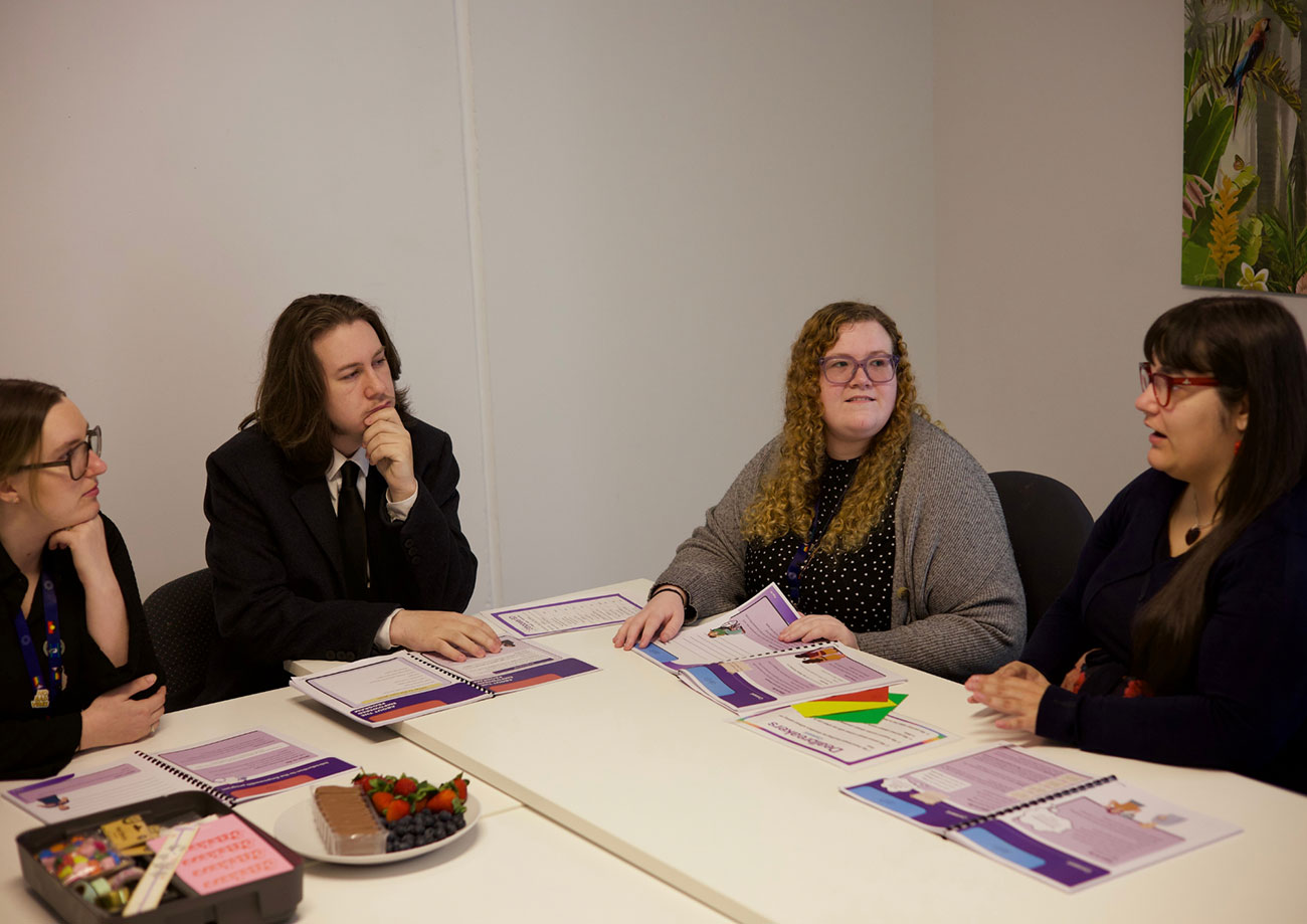 : Four team members sitting together listening to one of the team speaking, with documents and snacks on the table